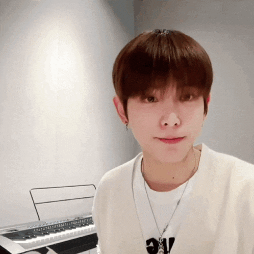 a young man wearing a white shirt and a necklace stands in front of a piano