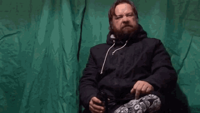 a man with a beard is sitting in front of a green cloth and giving the middle finger