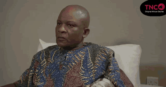 a bald man sitting on a bed with a tnc logo in the background