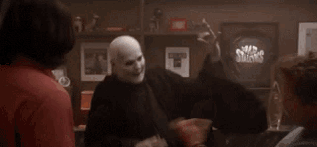 a bald man wearing a mask is standing in a room with other people and smoking a cigarette .