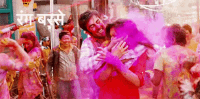 a man is holding a woman in his arms while a crowd of people are playing with colored powder .