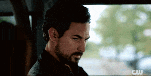 a man with a beard is sitting in a car with a cw logo behind him