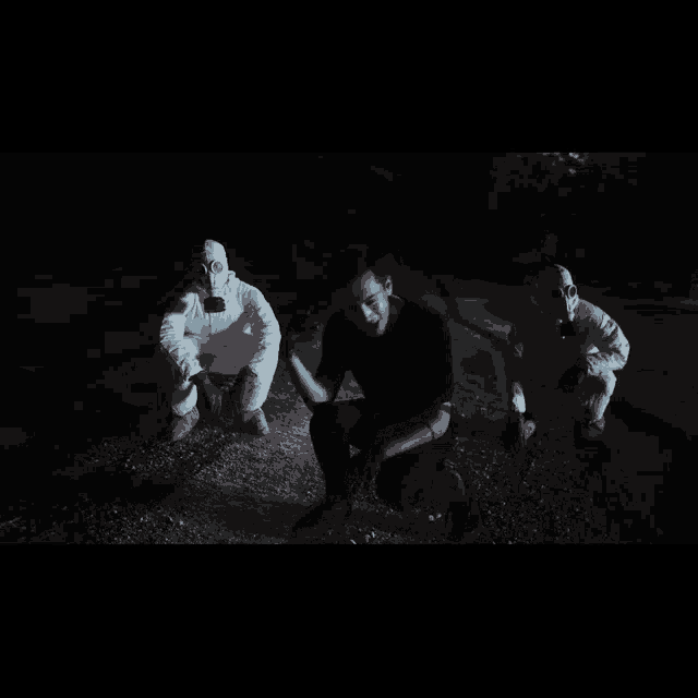 a man in a black shirt is kneeling down in front of two men in white gas masks