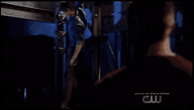 a man in a red shirt is standing in a dark room with a cw logo on the bottom of the screen