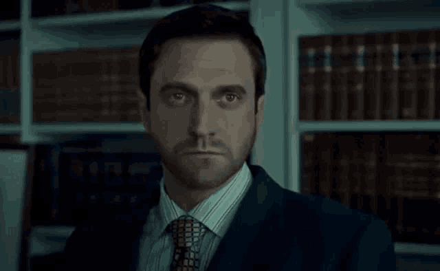 a man in a suit and tie stands in front of a book shelf