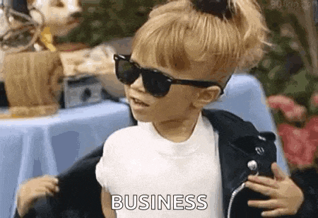 a little girl wearing sunglasses and a leather jacket is standing in front of a table and says `` business '' .