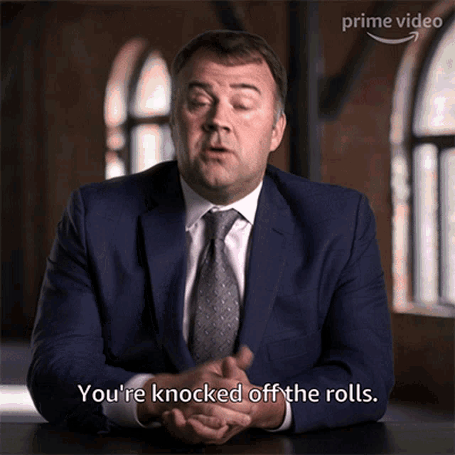 a man in a suit and tie is sitting at a table and says you 're knocked off the rolls