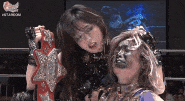 a female wrestler holds a stardom championship belt over another woman 's head