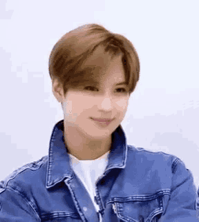 a young man wearing a blue denim jacket and a white shirt is looking at the camera .