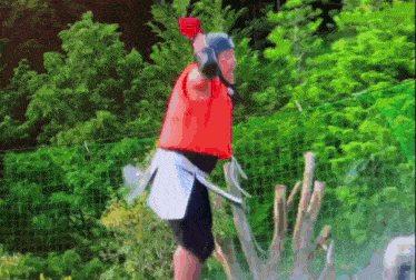 a man in a red vest and black shorts is standing in front of a fence holding a gun .