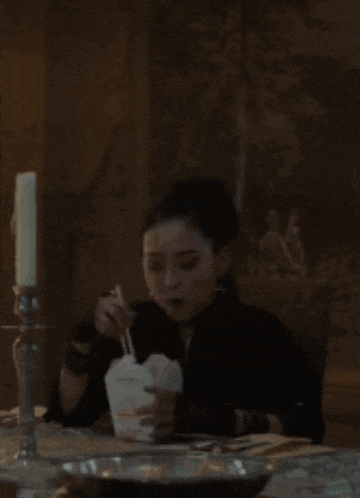 a woman is sitting at a table eating noodles with chopsticks from a takeout box