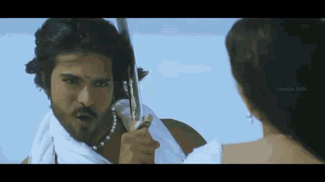 a man is holding a sword in front of a woman and the words geetha arts are on the bottom right