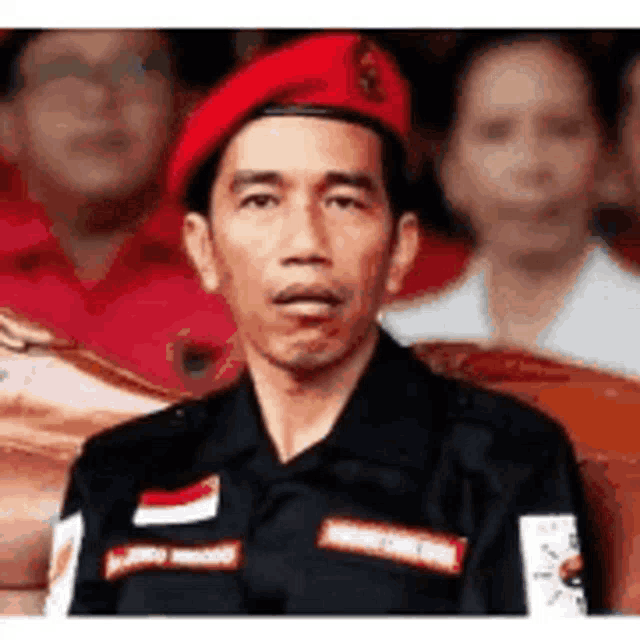 a man wearing a red beret and a black shirt is sitting in front of a crowd .
