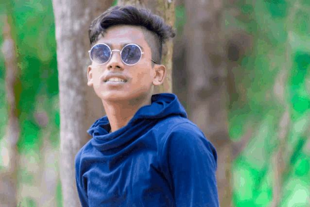 a young man wearing sunglasses and a blue shirt stands in front of a tree