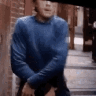a man in a blue sweater is standing in front of a brick wall with his arms crossed .