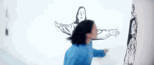 a woman in a blue jacket is standing in front of a drawing of a woman on a wall .