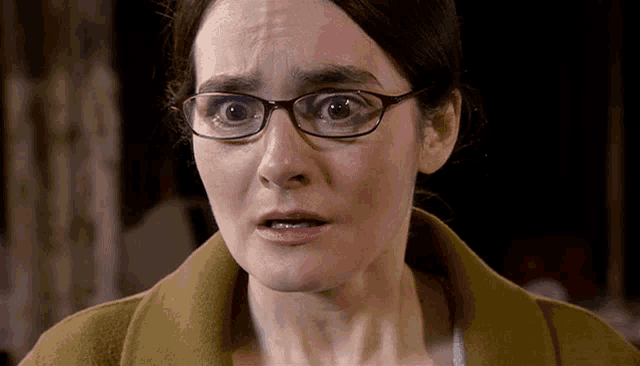 a woman wearing glasses and a brown coat is making a surprised face