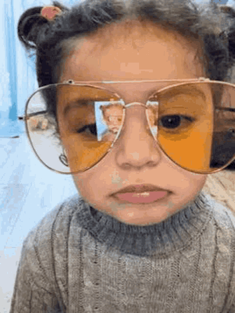 a little girl wearing sunglasses and a sweater is making a face .