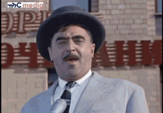 a man wearing a hat and tie is speaking into a microphone in front of a building that says mc media
