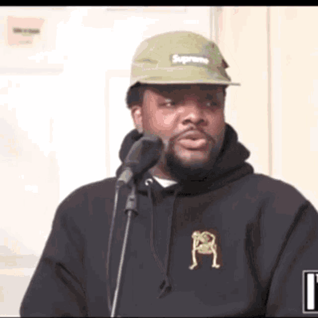 a man wearing a supreme hat and a black hoodie is standing in front of a microphone .