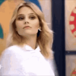 a woman with blonde hair is wearing a white hoodie and earrings and looking at the camera .