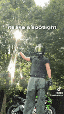 a man in a helmet stands in front of a motorcycle with the sun shining through the trees above him