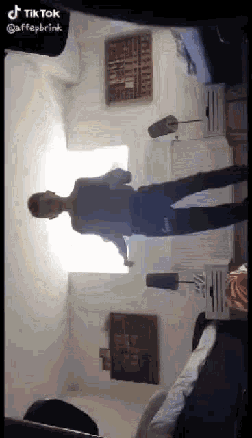 a man is standing in a room in front of a window with a lamp on the nightstand .