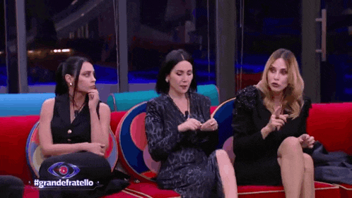 three women are sitting on a red couch with a sign that says grande fratello