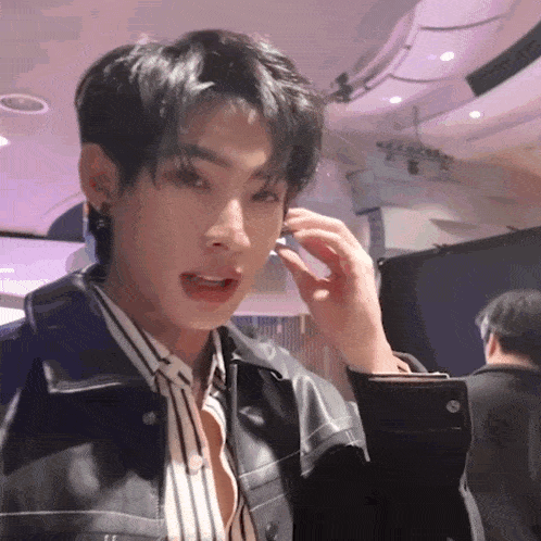 a young man adjusts his earring while wearing a striped shirt and a leather jacket