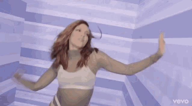 a woman in a white bra is dancing in front of a wall with the word vevo on the bottom
