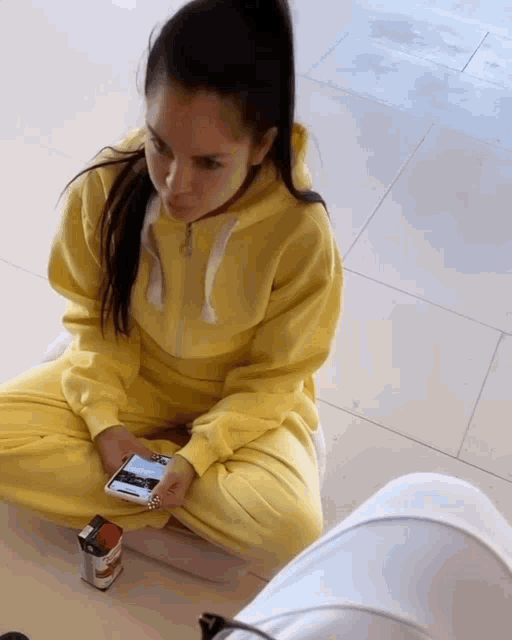 a woman in a yellow sweatshirt sits on the floor looking at her phone