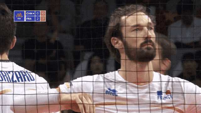 a man with a beard is behind a volleyball net and his jersey says brizard