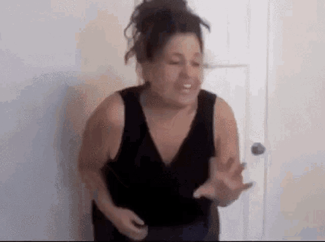 a woman in a black tank top is standing in front of a door and talking .