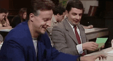 two men in suits and ties are sitting at a table in a classroom laughing .