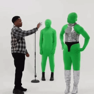 a man in a plaid shirt is standing in front of a microphone while two green aliens are standing in front of him .