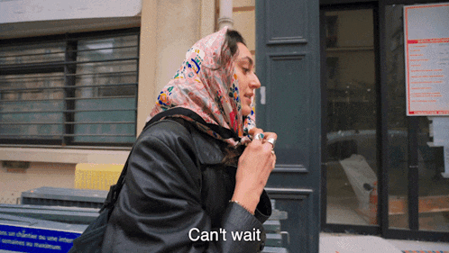 a woman with a scarf around her head says " can 't wait " in front of a building