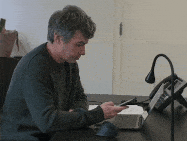 a man sits at a desk with a phone and a laptop looking at his phone