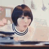 a woman is sitting at a table with a laptop computer and making a surprised face .