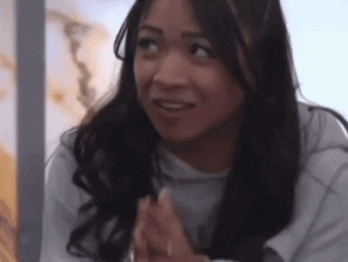 a woman with long dark hair is smiling with her hands folded in front of her face .