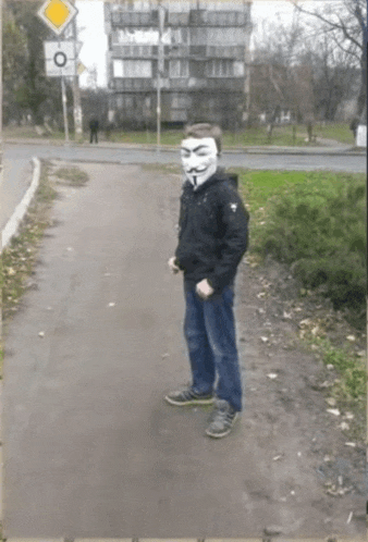 a man wearing an anonymous mask is standing on a sidewalk .