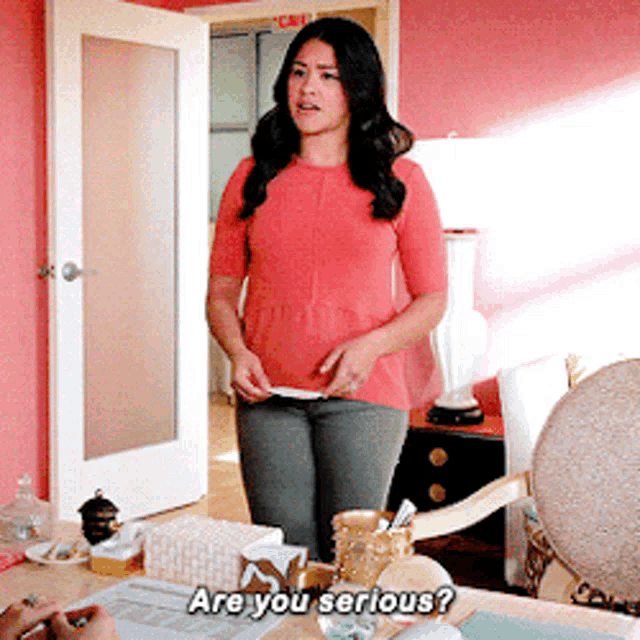 a woman in a red shirt is standing in front of a desk and says are you serious