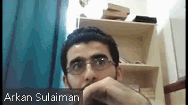 a man with glasses is sitting in front of a bookshelf with the name arkan sulaiman on the bottom right