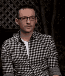 a man wearing glasses and a plaid shirt is sitting in front of a lattice wall .