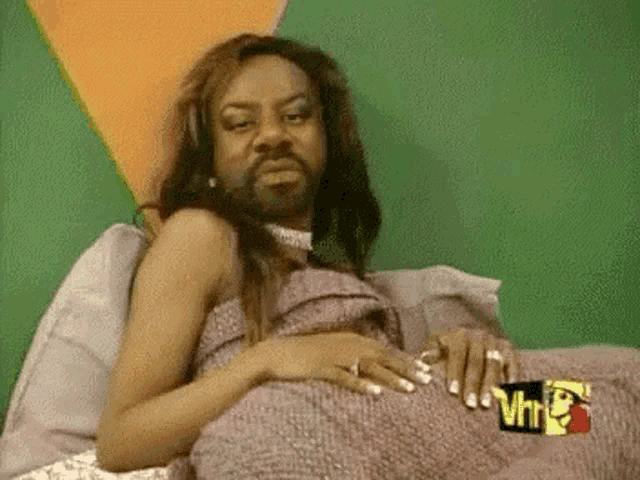 a woman with a beard is laying on a bed with a vh1 logo on the wall .