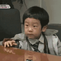 a young boy in a suit and tie is sitting at a table with his hand on the table .