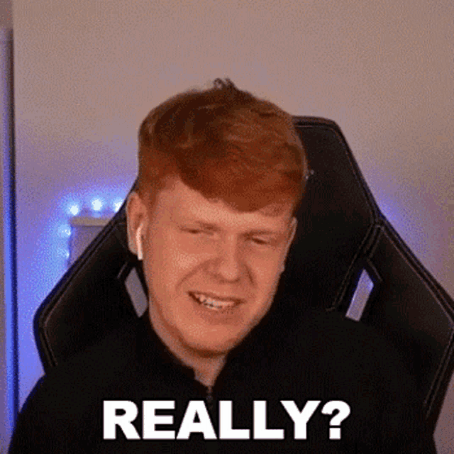 a man with red hair is sitting in a gaming chair and smiling while wearing headphones .