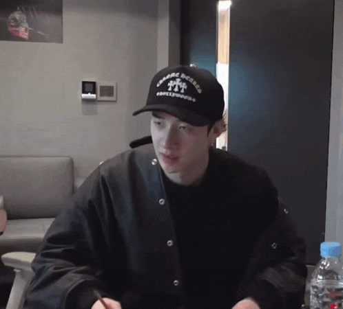 a young man wearing a black hat and a black jacket is sitting in a room .