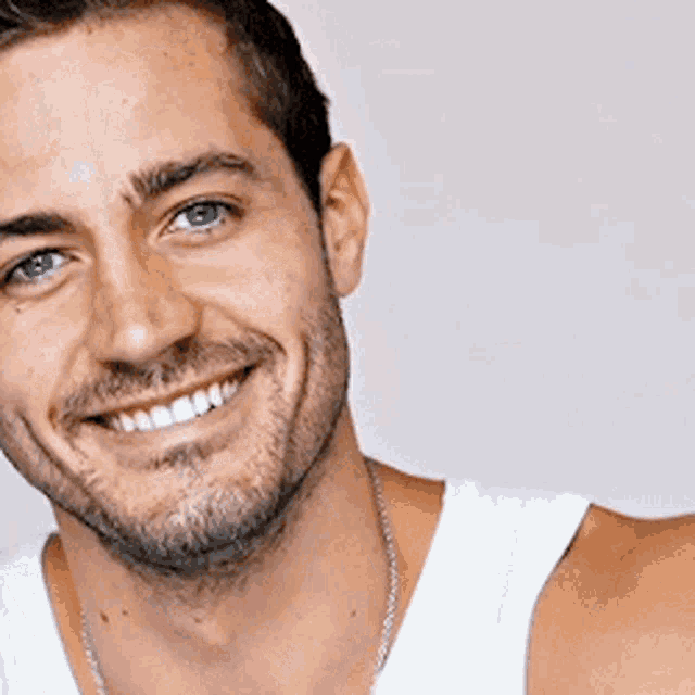 a man wearing a white tank top and a necklace smiles for the camera