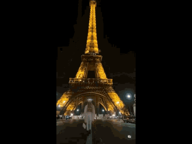 a woman in a white coat is walking in front of the eiffel tower at night .