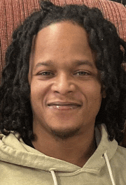 a man with dreadlocks is wearing a tan hoodie and smiling